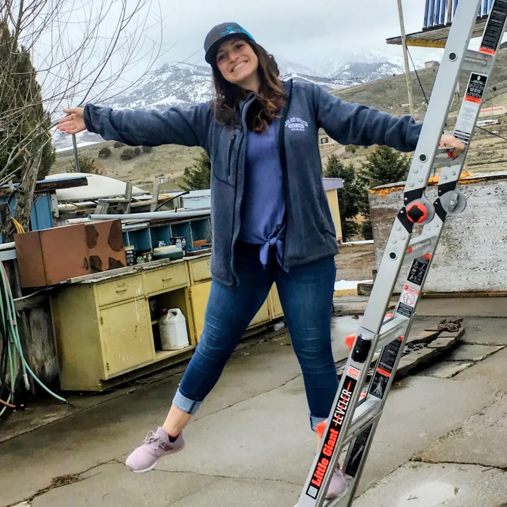 A Lavalink team member at a tower site above Lava Hot Springs, arms flung wide and one foot off the ladder, snow still on the mountains behind